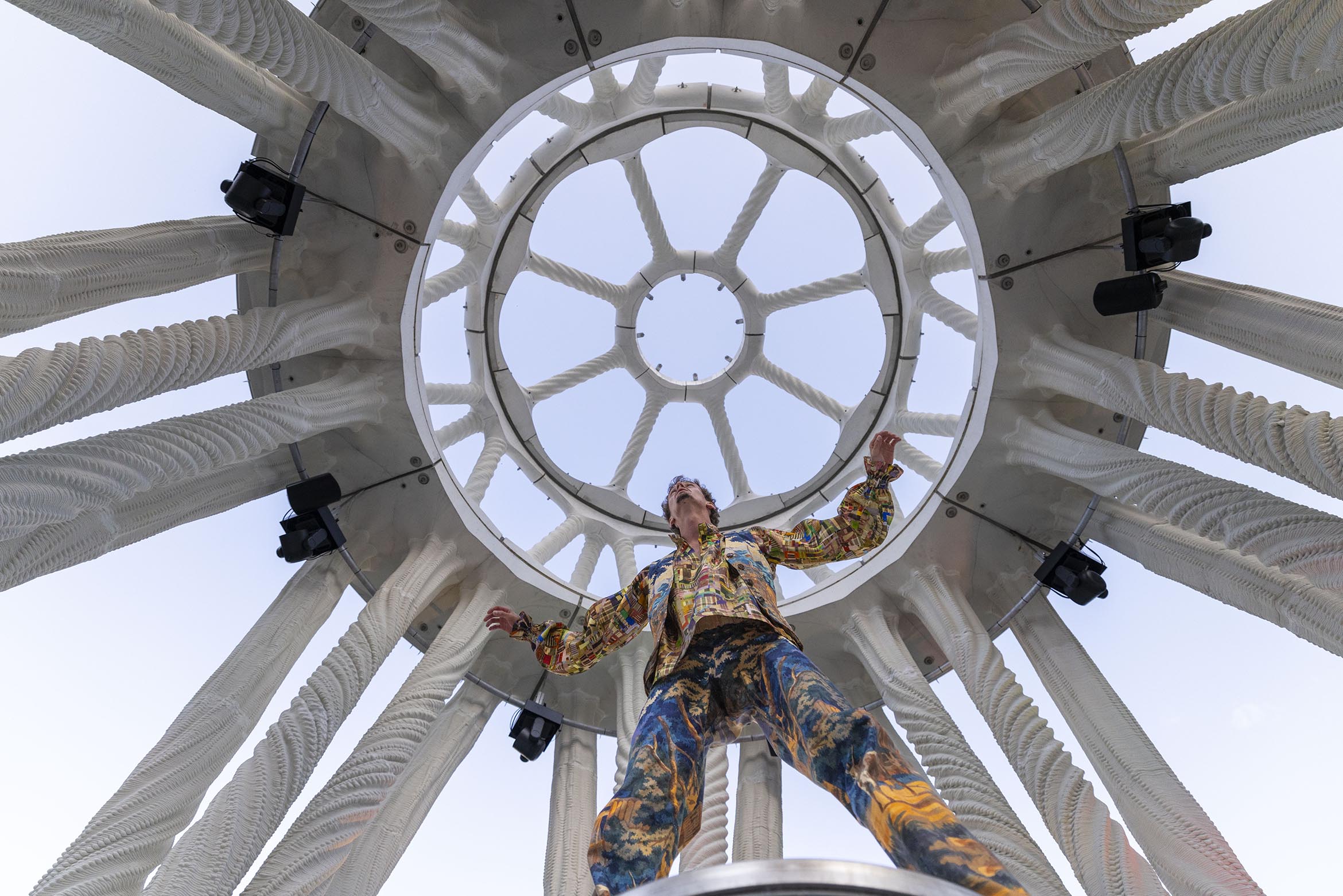 Person von unten fotografiert unter einer kreisförmigen architektonischen Struktur mit radial angeordneten Säulen, trägt farbige gemusterte Kleidung